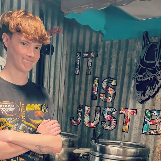A red-haired regular, arms crossed, in the shed. Corrugated-metal walls and a sticker-collage sign reading It Is Just Beer behind him.