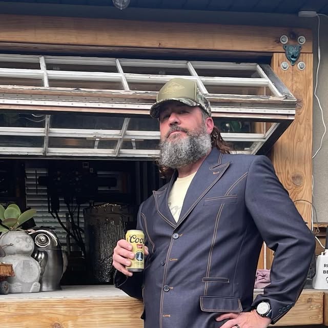 Steve Birch, bearded, in a camo cap and a sharp blazer, holding a can of Coors in front of the Brewshed.